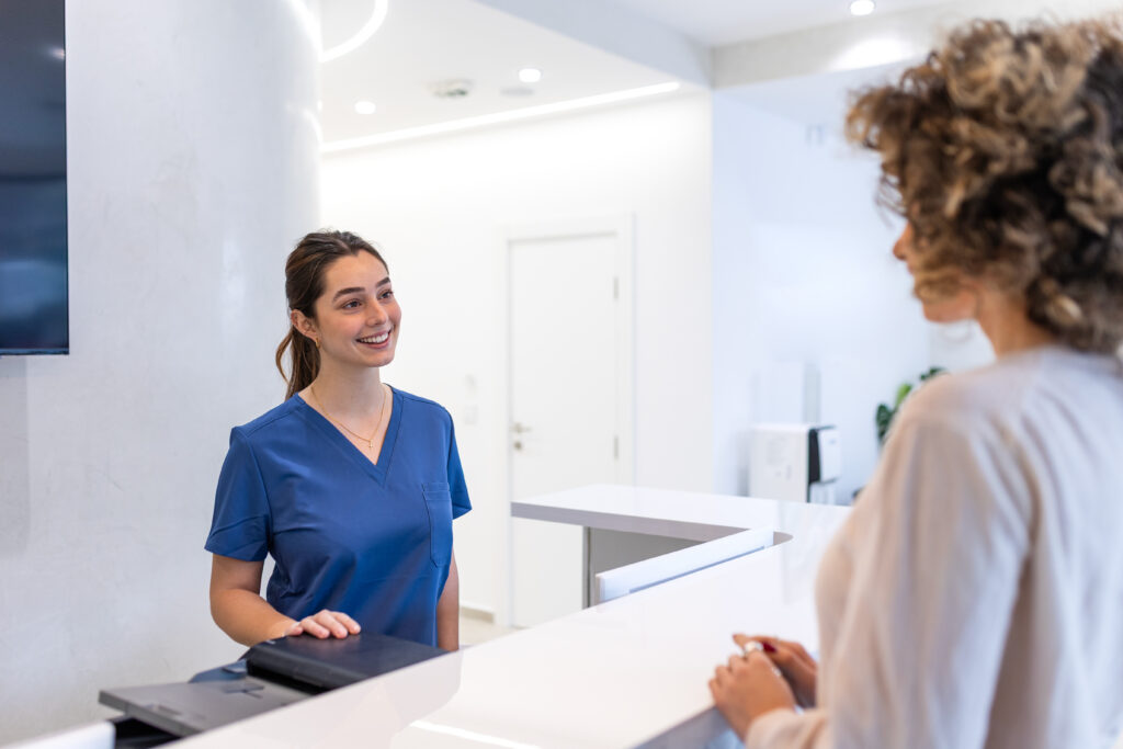 Friendly Medical Receptionist Welcoming Patient at Hospital Front Desk. Smiling Professional in Blue Scrub Uniform at Modern Clinic Reception Area, Healthcare Service and Consultation Concept.