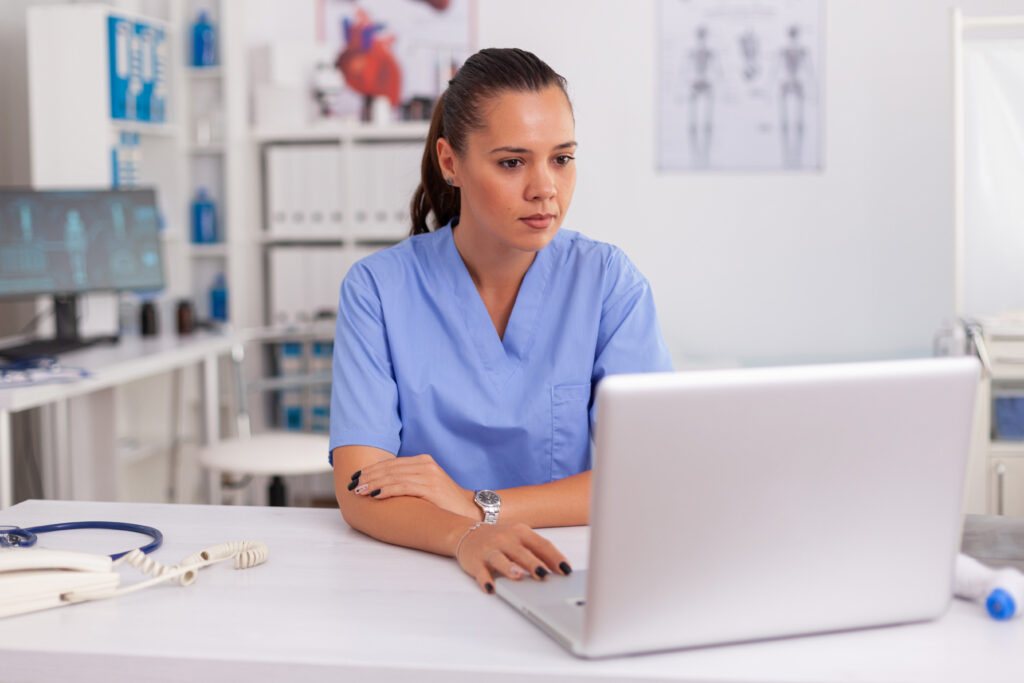 How AI Clinical Documentation Supports Financial Workflows Medical nurse working on laptop in hospital office wearing blue uniform. Health care practitioner sitting at desk using computer in modern clinic looking at monitor, medicine.