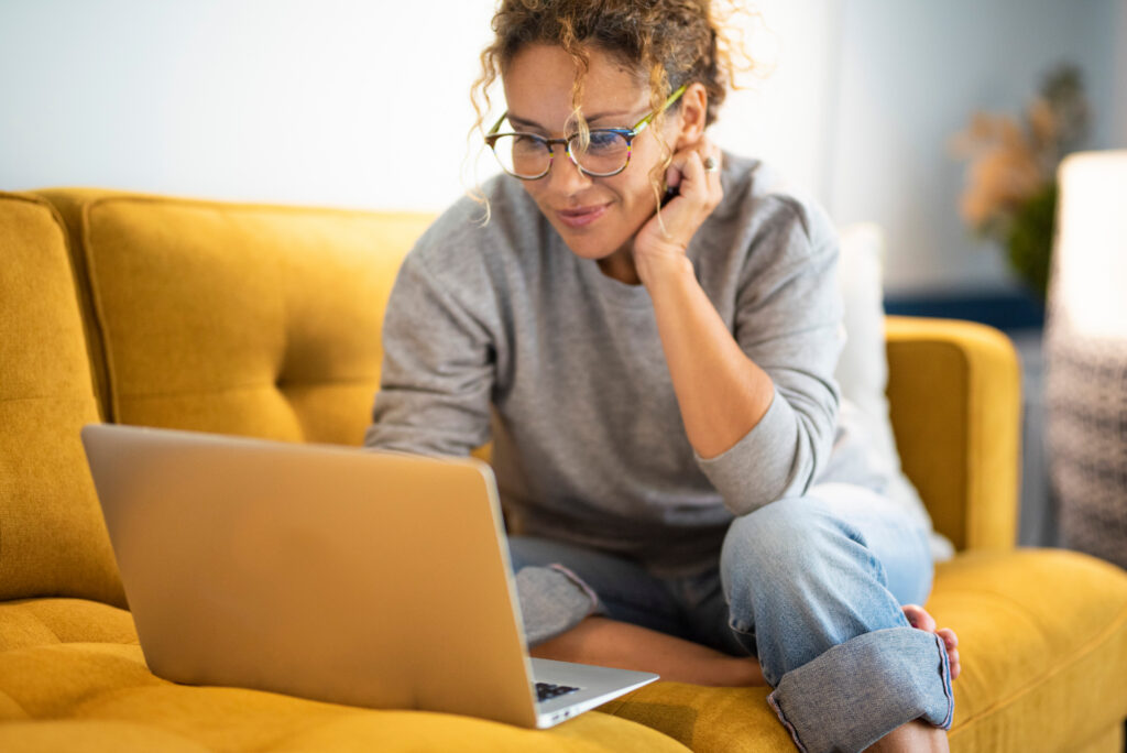 Creating a Connected Patient Scheduling Experience Across Web, Mobile, and Search Female sitting on a yellow couch using laptop and internet connection and smile. Happy woman in indoor technology leisure activity. Social media life account concept. Adult Lady writing on computer