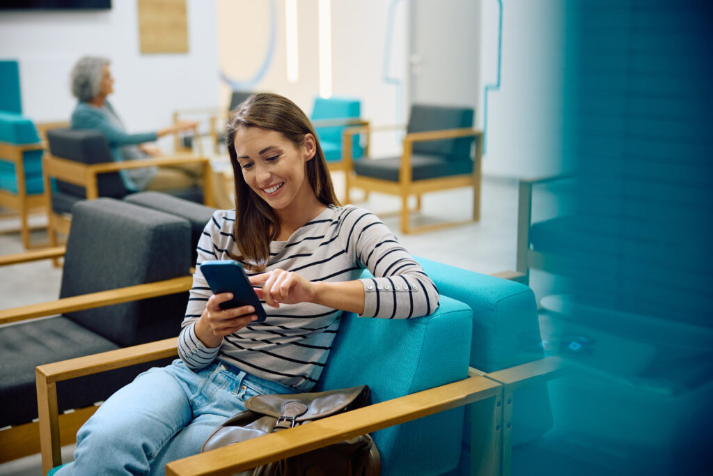 Creating a Connected Patient Scheduling Experience Across Web, Mobile, and Search Young happy woman using cell phone in waiting room at doctor's office.