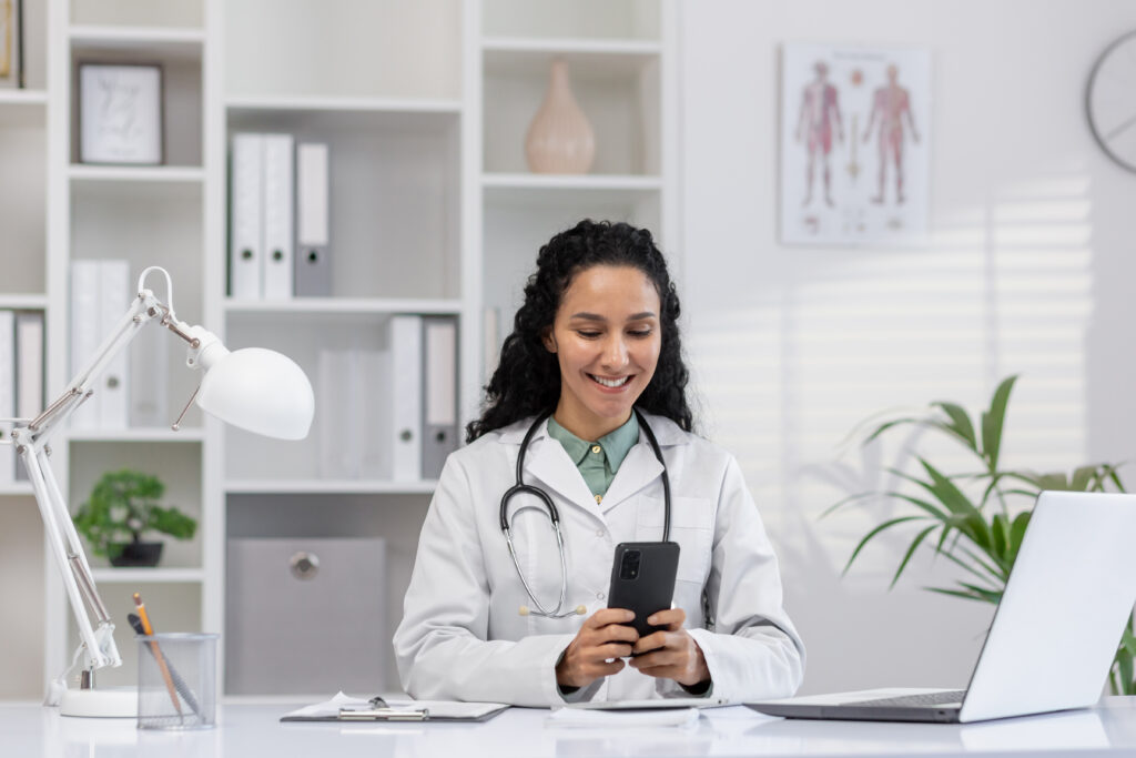Why Workflow Simplicity Is Vital for Independent Practices Cheerful female doctor in a white coat using a smartphone in a modern clinic office. She is smiling and appears engaged and relaxed in her professional environment.