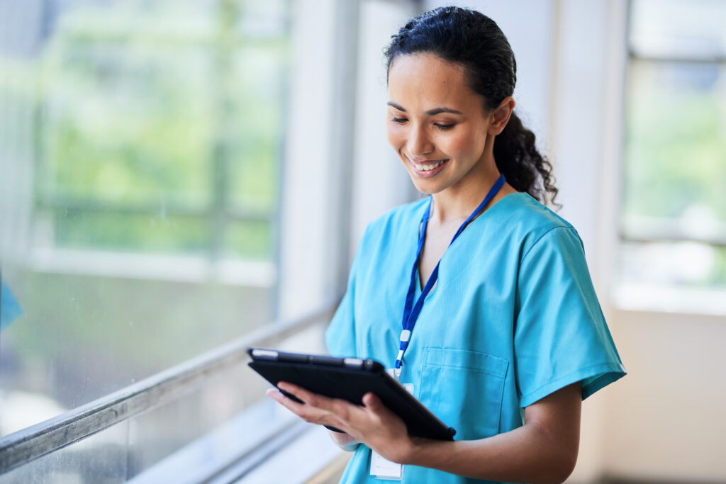 Why Front-Office Patient Communication Workflows Matter Smiling nurse wearing hospital scrubs and holding a digital tablet while standing in a modern medical facility. The image portrays a professional medical worker in a healthcare environment, using technology for patient care.
