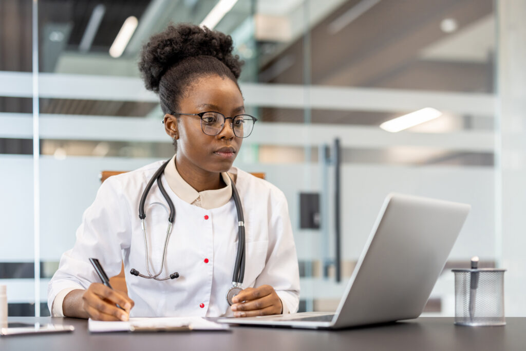 Why AI Clinical Documentation Software Is the New Standard Doctor diligently working on laptop and writing notes in a modern medical office. Professional healthcare provider wearing a stethoscope, demonstrating concentration and dedication to her work.